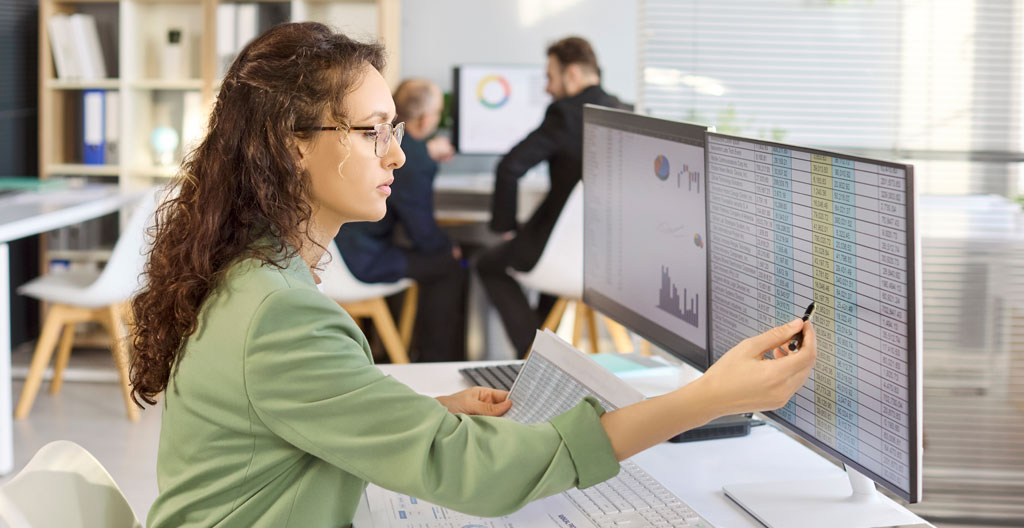 Artisan plombier dans son atelier professionnel, debout devant un établi métallique, consultant un document URSSAF ou une facture d'assurance, environnement de travail avec tuyauterie et outillage visible